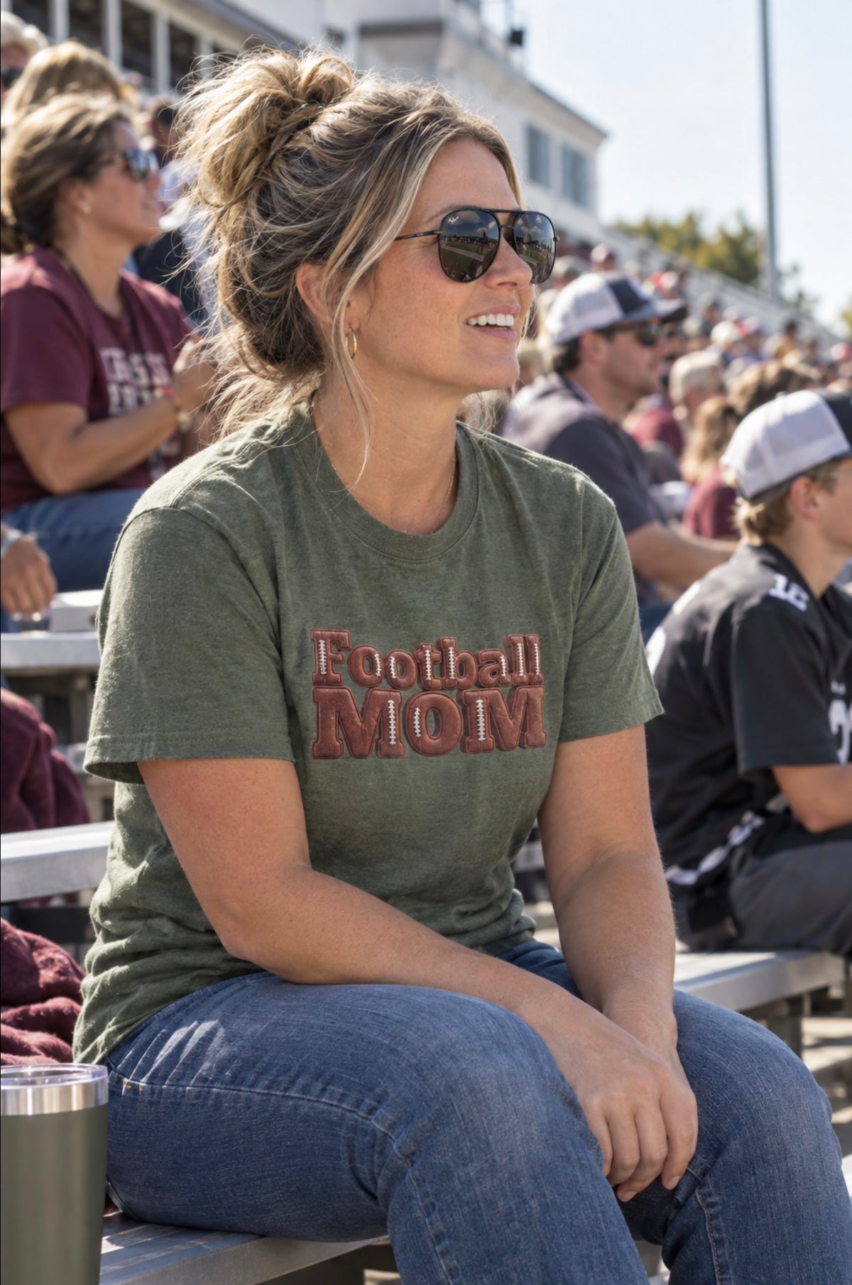 A mom sitting in stadium bleachers wearing an military green graphic t-shirt that reads 'Football MOM' in a 3D block font filled with a brown football leather texture and white laces.