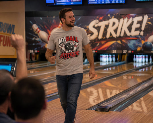 A man in a bowling alley wearing a light gray graphic t-shirt that reads 'MY BALL HAS ATTITUDE' and dark blue jeans, walking down the lane after a game. The background shows bowling pins, lanes, and a colorful 'STRIKE!' sign.