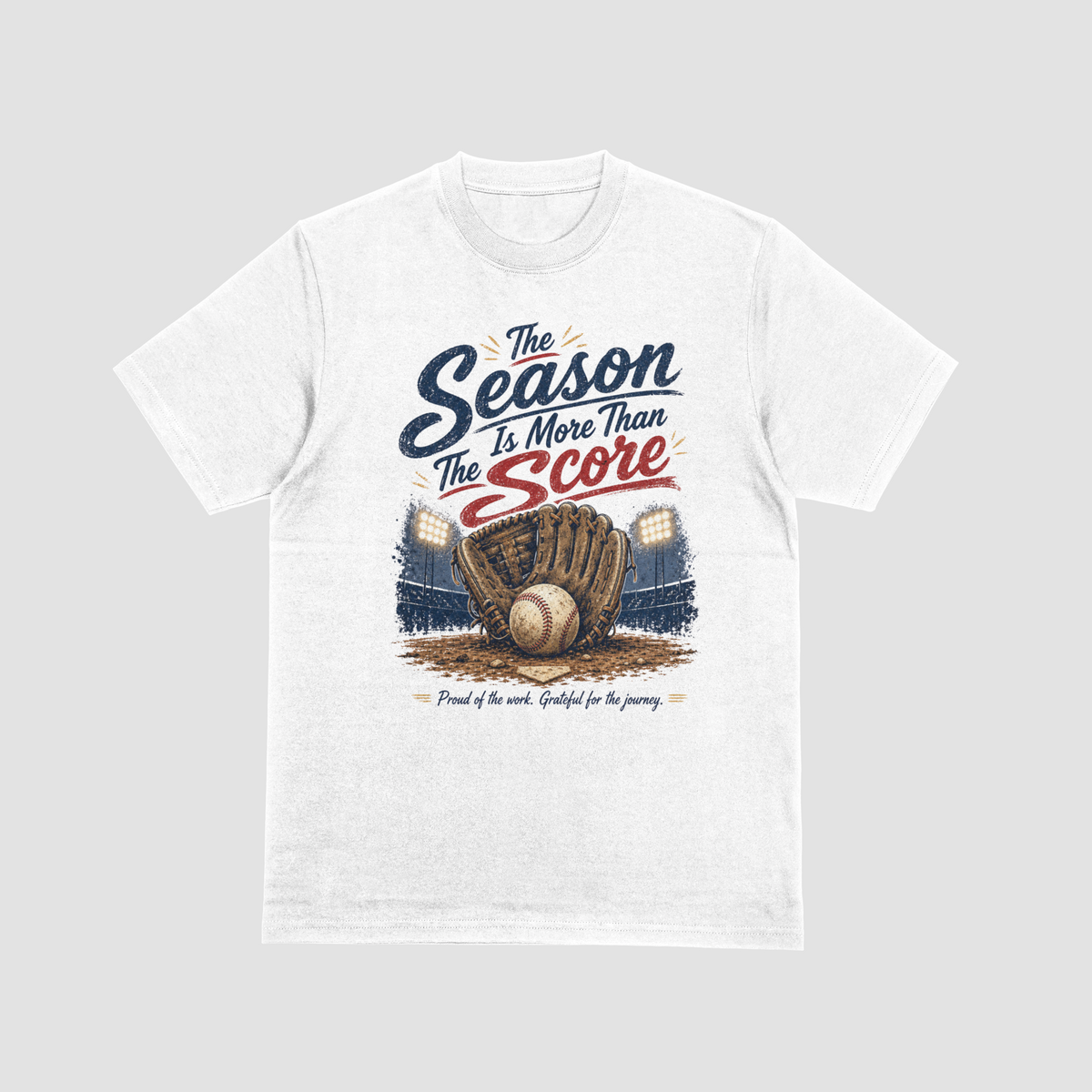 A white short-sleeve crewneck t-shirt featuring a vintage-style baseball graphic. The design reads “The Season Is More Than The Score” in navy blue and red lettering above a brown leather baseball glove holding a baseball, with a baseball field and illuminated stadium lights in the background. The design also includes the phrase “Proud of the work. Grateful for the journey.”