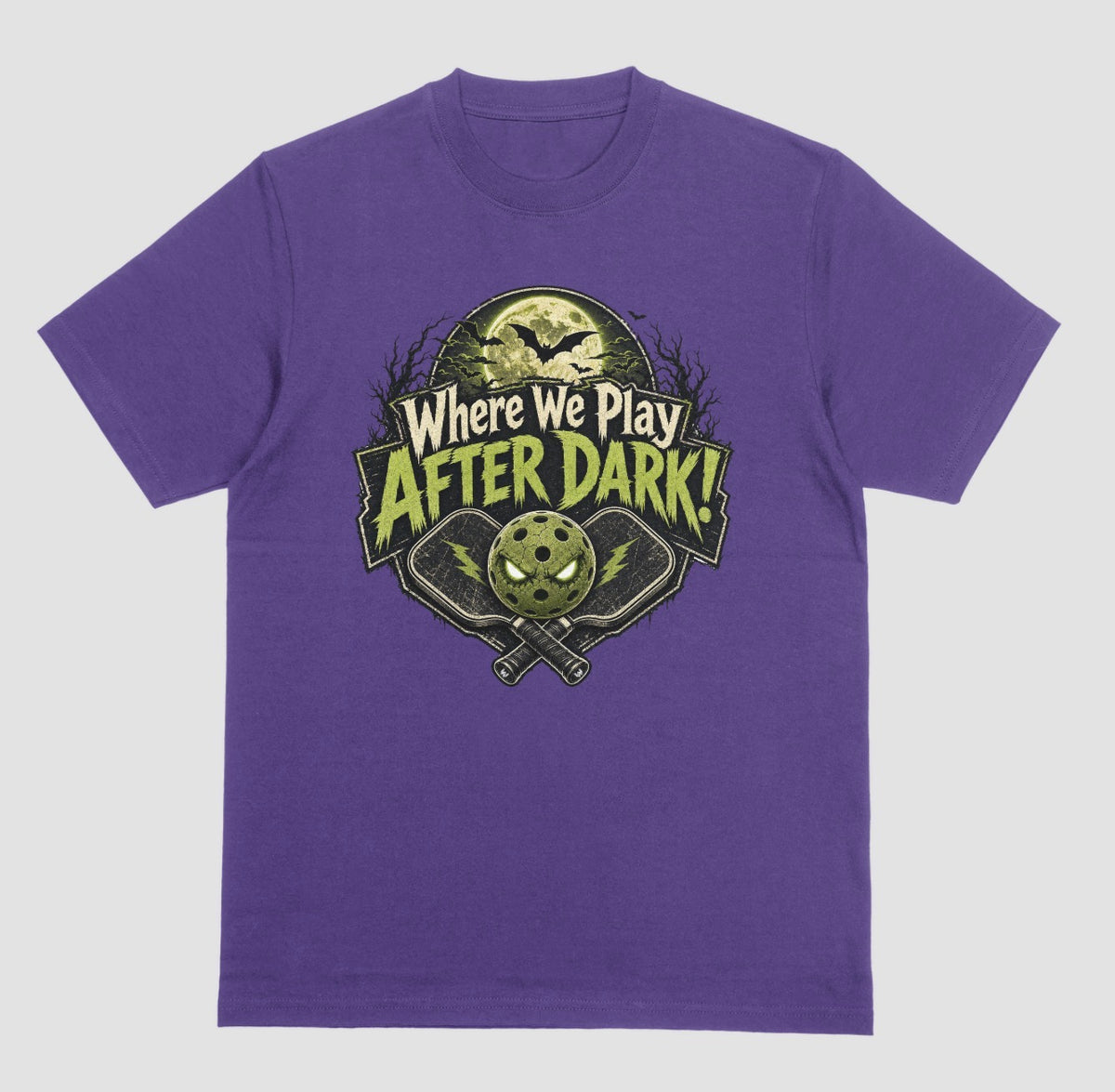 A purple t-shirt with a spooky pickleball graphic reading 'Where We Play After Dark!' showing crossed paddles and a glowing-eyed pickleball under a full moon with bats.
