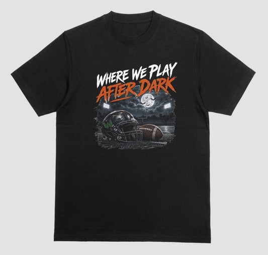 A black short-sleeve graphic t-shirt featuring the text 'WHERE WE PLAY AFTER DARK' in white and orange lettering above a football-themed illustration, displayed on a neutral light gray background.