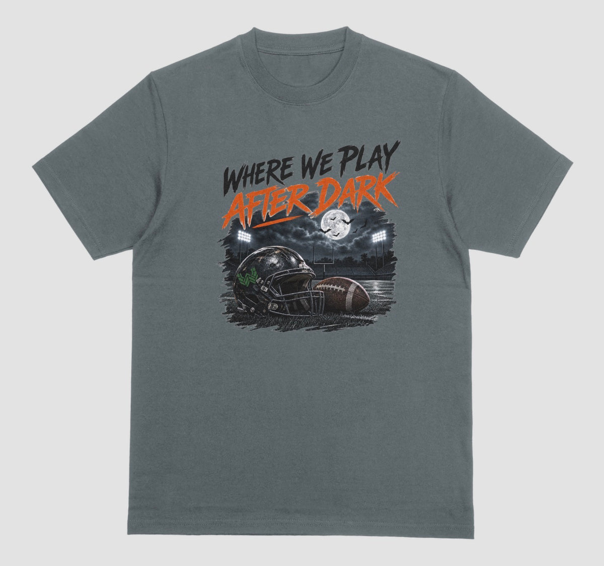 A dark heather crewneck t-shirt featuring a graphic print on the front with the text 'WHERE WE PLAY AFTER DARK' in black and orange, above an illustration of a football helmet and football in a stadium at night.