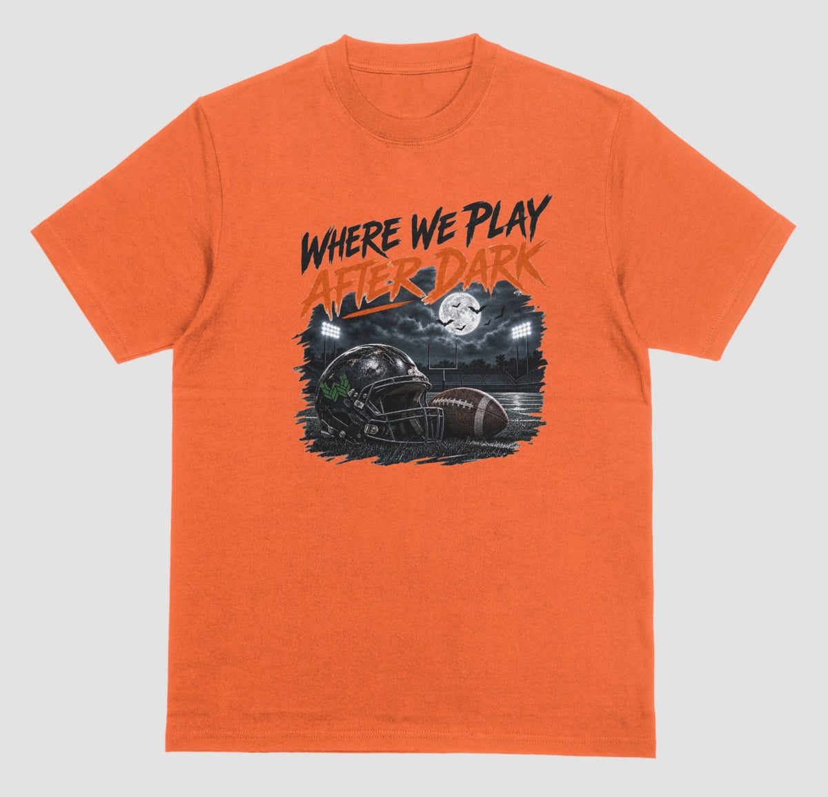 An orange crewneck t-shirt featuring a graphic print on the front with the text 'WHERE WE PLAY AFTER DARK' in black and orange, above an illustration of a football helmet and football in a stadium at night.