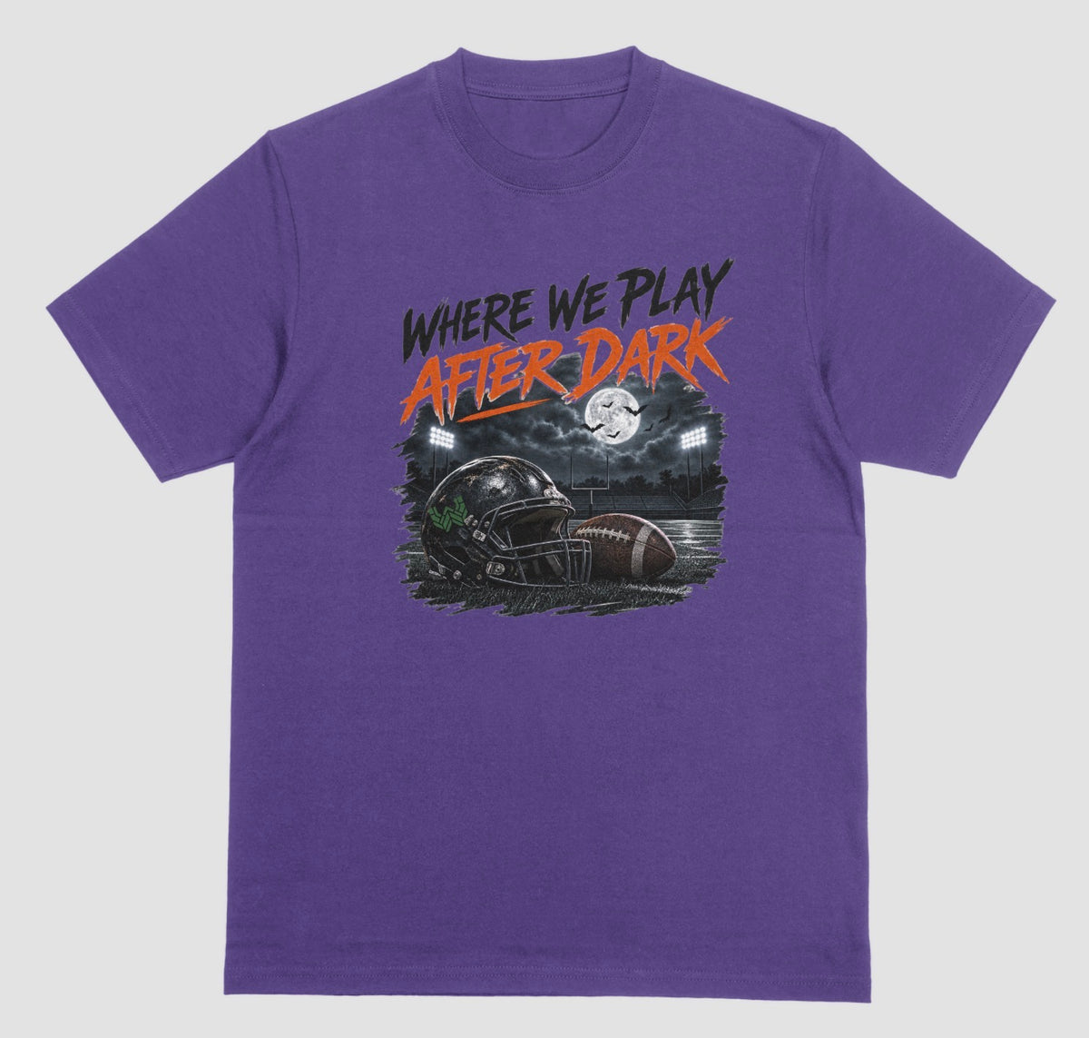 A purple crewneck t-shirt featuring a graphic print on the front with the text 'WHERE WE PLAY AFTER DARK' in black and orange, above an illustration of a football helmet and football in a stadium at night.