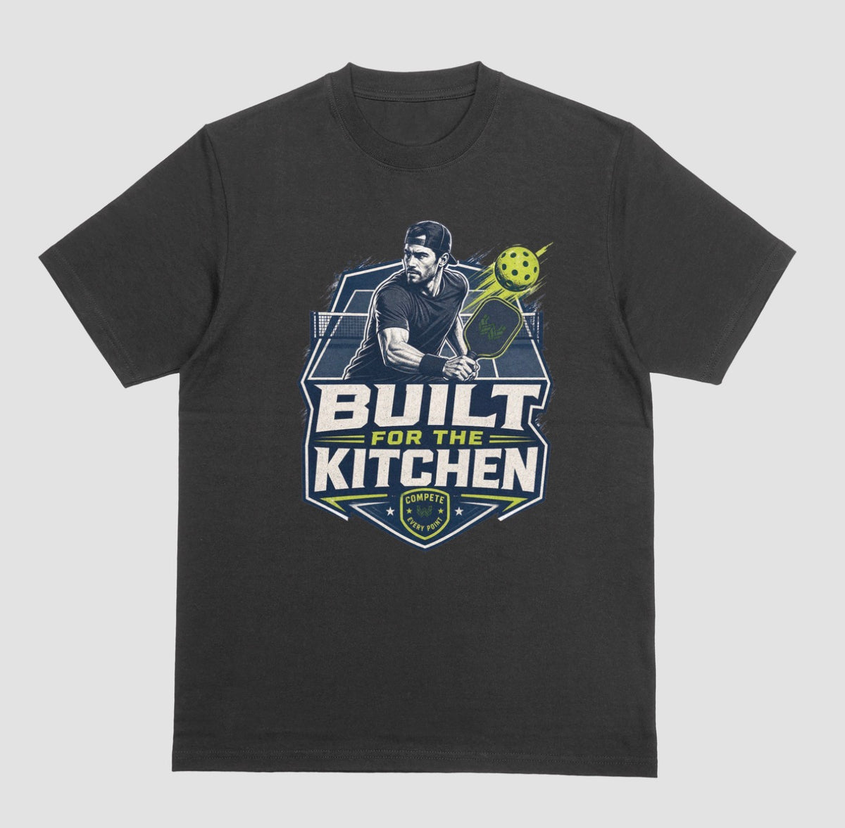 A black crewneck t-shirt featuring a large graphic print on the front. The graphic depicts a man holding a pickleball paddle with a yellow-green pickleball, above the text 'BUILT FOR THE KITCHEN' in white and yellow, with a 'SPORTS' shield logo at the bottom.