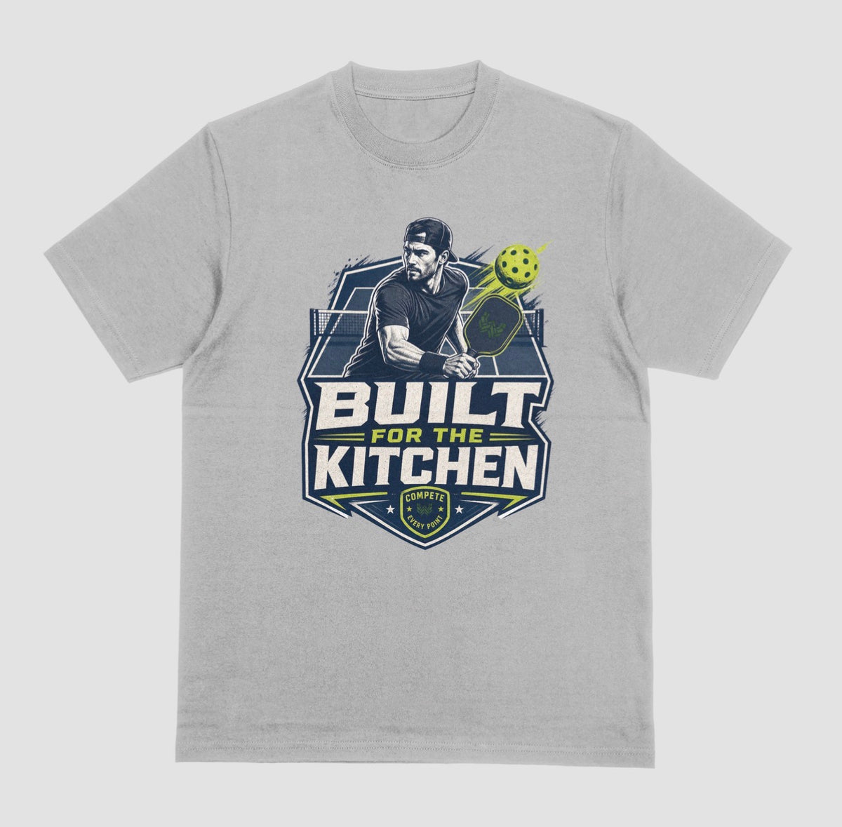 A gray crewneck t-shirt featuring a large graphic print on the front. The graphic depicts a man holding a pickleball paddle with a yellow-green pickleball, above the text 'BUILT FOR THE KITCHEN' in white and yellow, with a 'SPORTS' shield logo at the bottom.