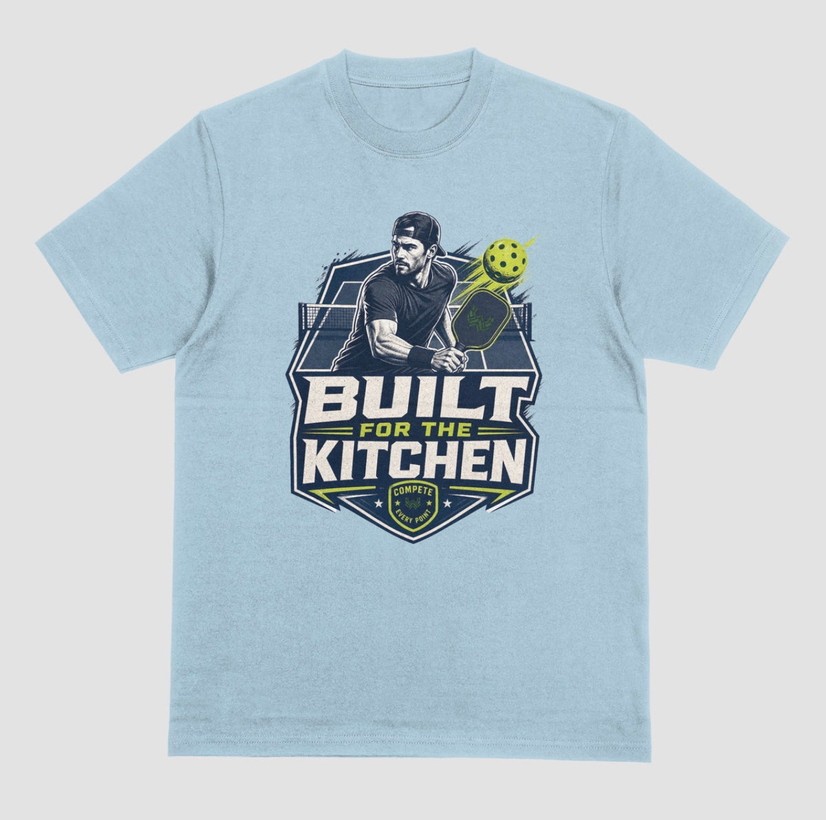 I light blue crewneck t-shirt featuring a large graphic print on the front. The graphic depicts a man holding a pickleball paddle with a yellow-green pickleball, above the text 'BUILT FOR THE KITCHEN' in white and yellow, with a 'SPORTS' shield logo at the bottom.