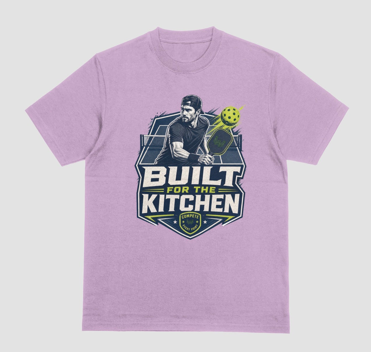 A lilac crewneck t-shirt featuring a large graphic print on the front. The graphic depicts a man holding a pickleball paddle with a yellow-green pickleball, above the text 'BUILT FOR THE KITCHEN' in white and yellow, with a 'SPORTS' shield logo at the bottom.