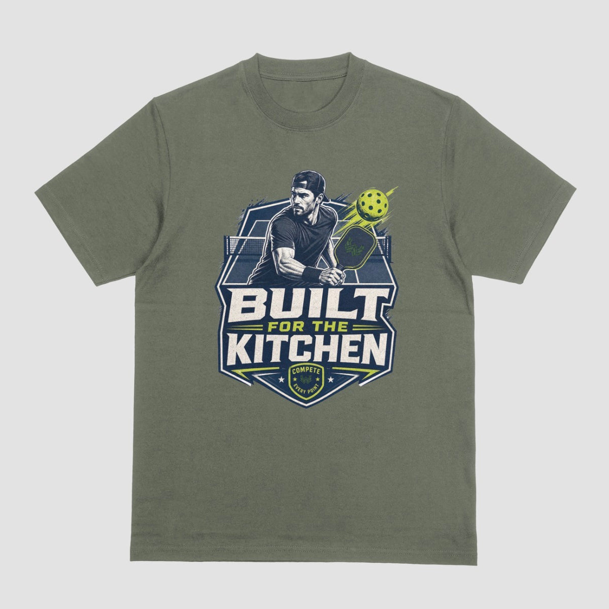 A military green crewneck t-shirt featuring a large graphic print on the front. The graphic depicts a man holding a pickleball paddle with a yellow-green pickleball, above the text 'BUILT FOR THE KITCHEN' in white and yellow, with a 'SPORTS' shield logo at the bottom.