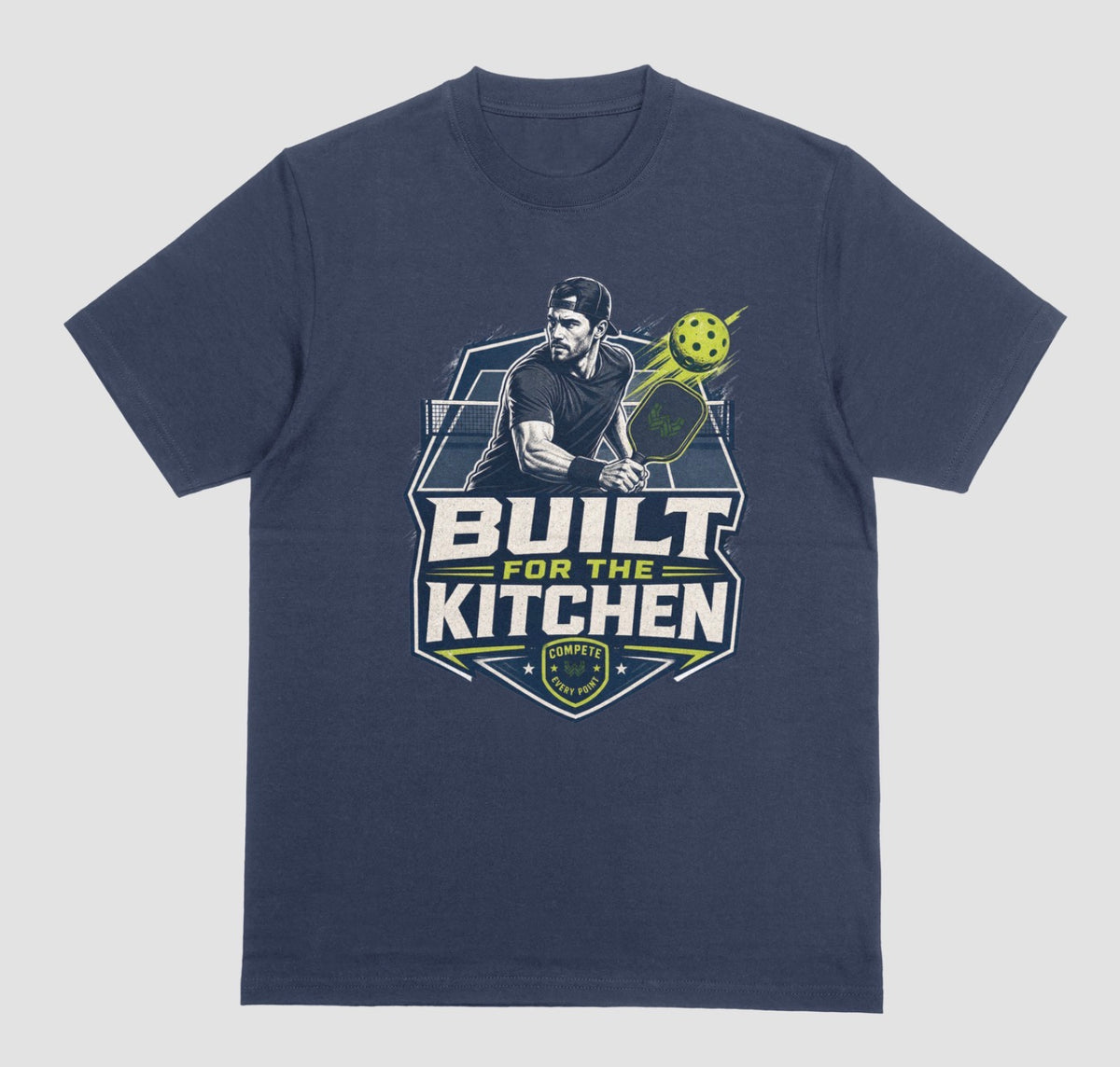A navy blue crewneck t-shirt featuring a large graphic print on the front. The graphic depicts a man holding a pickleball paddle with a yellow-green pickleball, above the text 'BUILT FOR THE KITCHEN' in white and yellow, with a 'SPORTS' shield logo at the bottom.