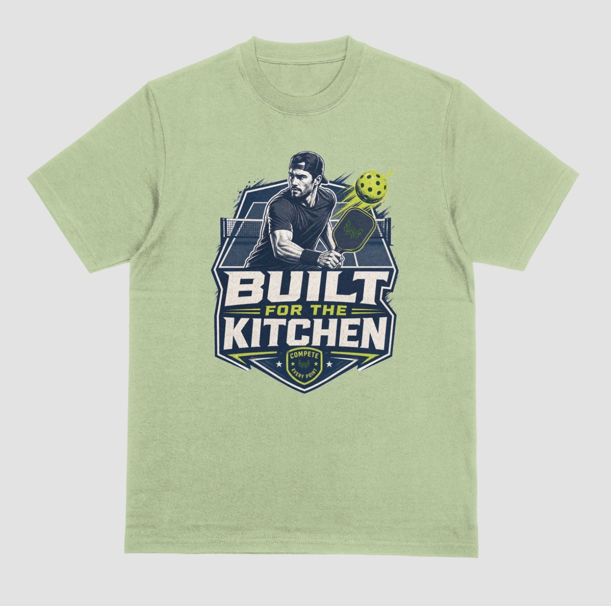 A pistachio crewneck t-shirt featuring a large graphic print on the front. The graphic depicts a man holding a pickleball paddle with a yellow-green pickleball, above the text 'BUILT FOR THE KITCHEN' in white and yellow, with a 'SPORTS' shield logo at the bottom.