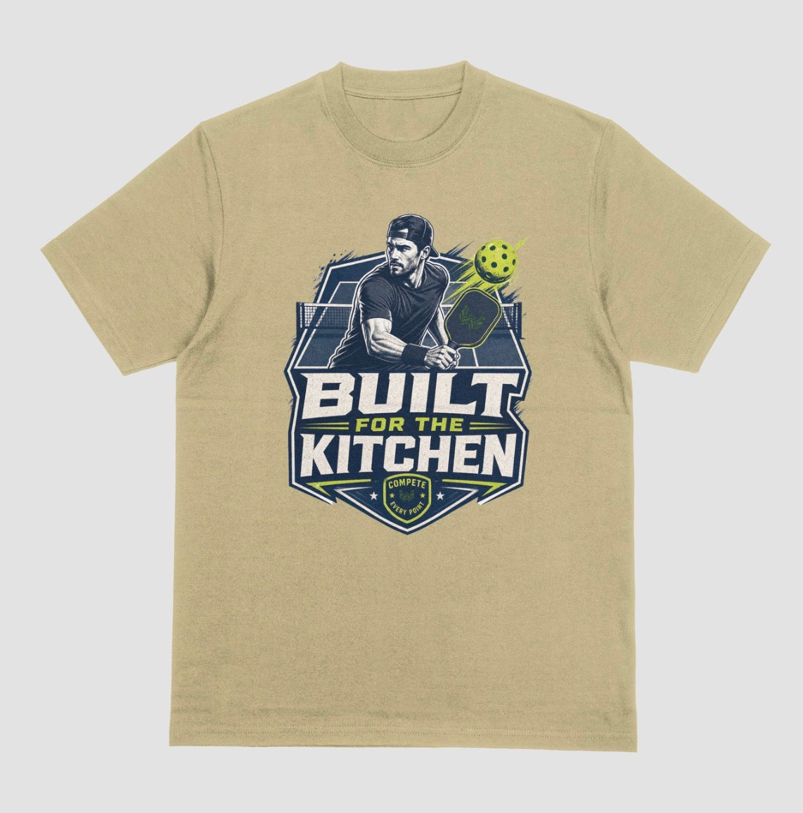 A sand color crewneck t-shirt featuring a large graphic print on the front. The graphic depicts a man holding a pickleball paddle with a yellow-green pickleball, above the text 'BUILT FOR THE KITCHEN' in white and yellow, with a 'SPORTS' shield logo at the bottom.
