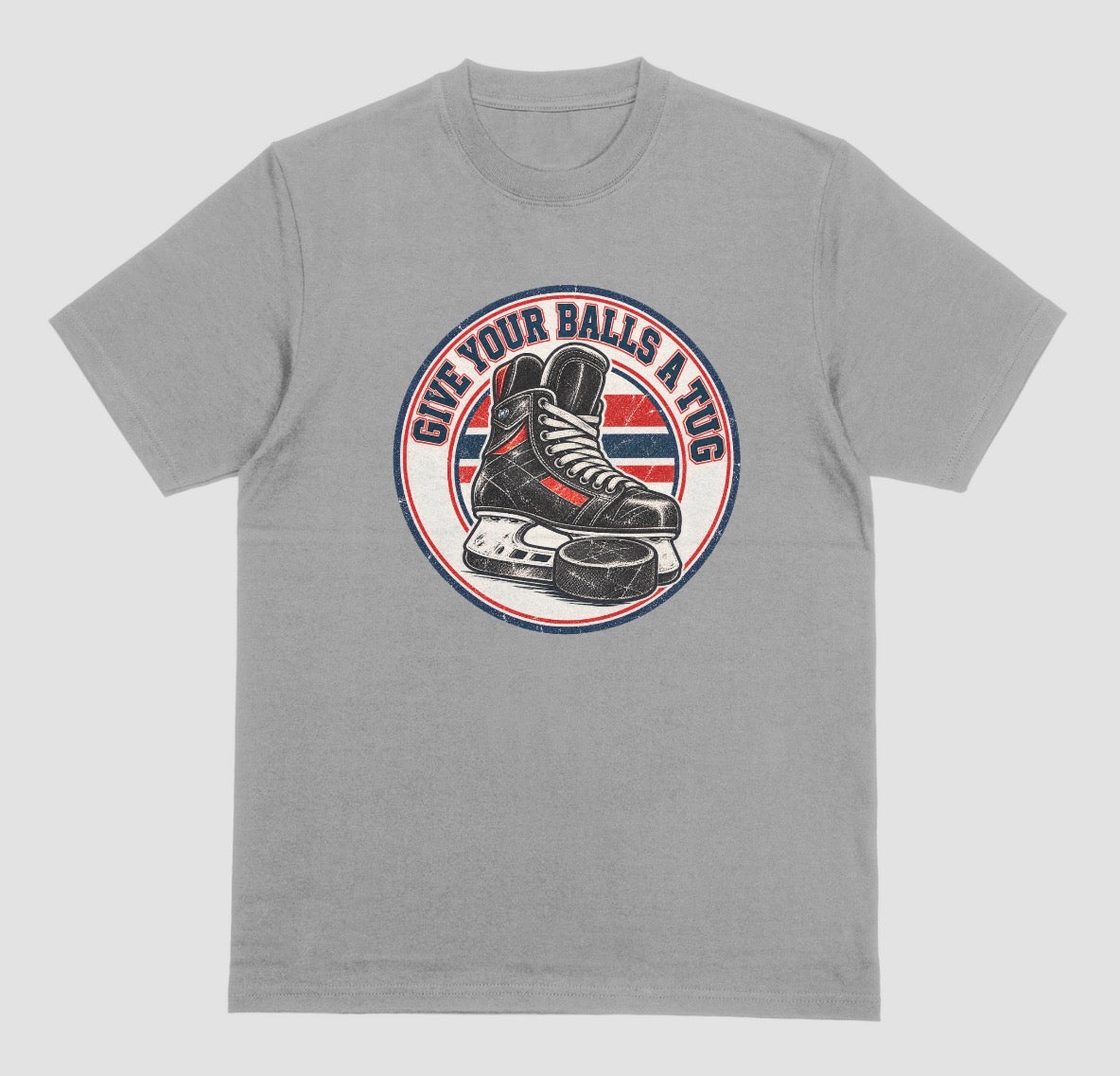 A grey t-shirt with a round vintage crest reading 'Give Your Balls a Tug' around a hockey skate and puck in red, white, and blue.