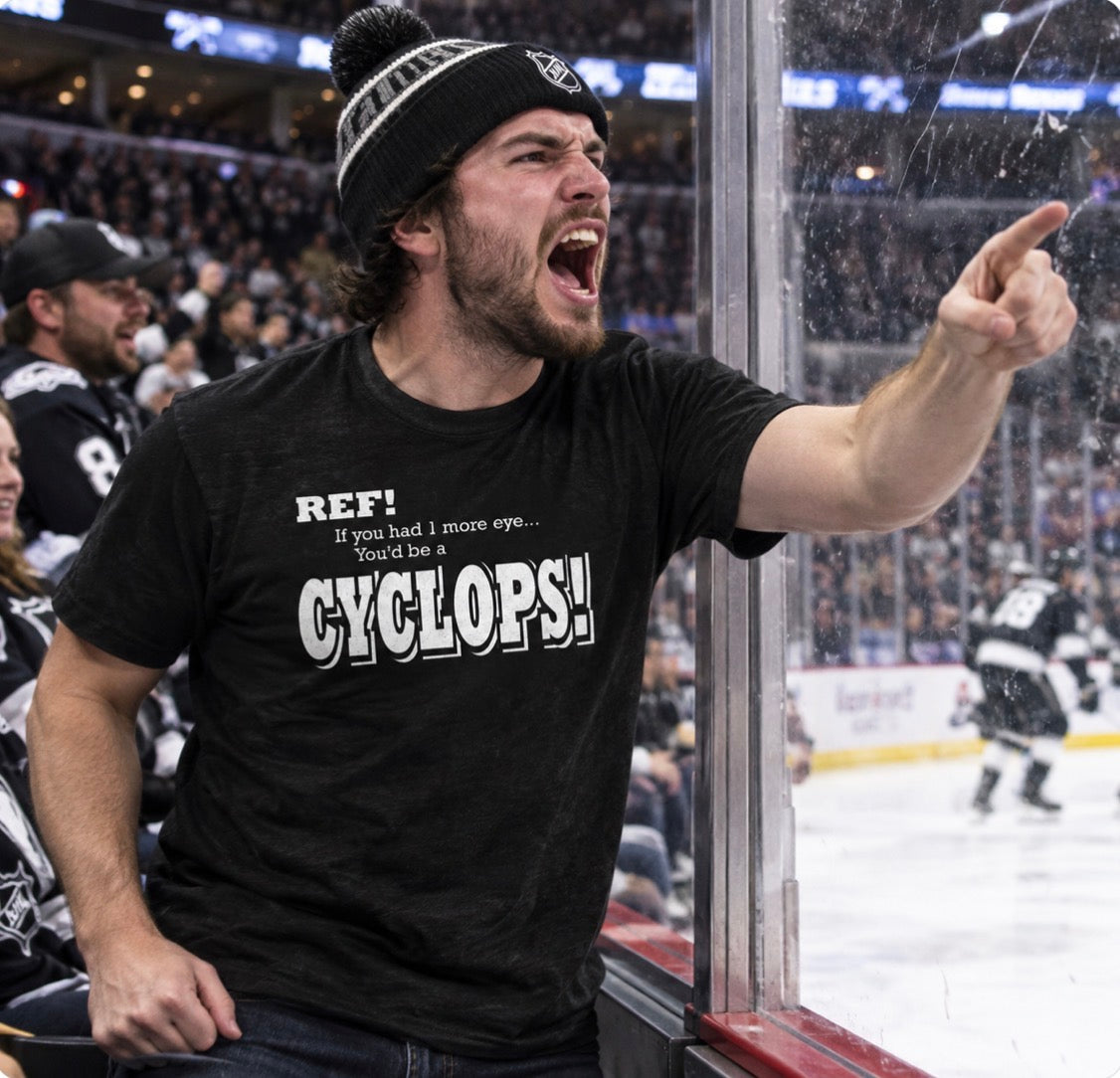 Man in a black beanie and black "Ref! If you had 1 more eye... You'd be a Cyclops!" t-shirt yelling and pointing from the stands at a hockey game