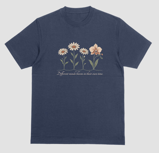 A navy blue t-shirt with an illustration of three daisies and an orchid and the script text ‘Different minds bloom in their own time.’