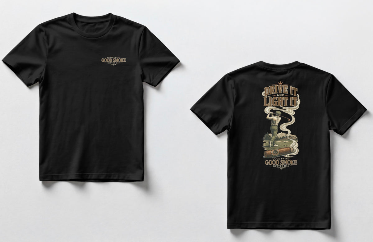 A black t-shirt, front and back views. Front chest features small text reading 'Good Swing, Good Smoke, Better Day.' Back features a large vintage-style illustration of a golfer mid-swing on a course, with a cigar in the foreground releasing swirling smoke that wraps around the text 'Drive It and Light It' at the top, with 'Good Swing, Good Smoke, Better Day' printed below the graphic.