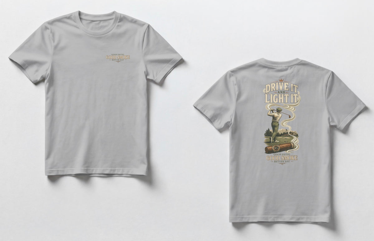 A grey t-shirt, front and back views. Front chest features small text reading 'Good Swing, Good Smoke, Better Day.' Back features a large vintage-style illustration of a golfer mid-swing on a course, with a cigar in the foreground releasing swirling smoke that wraps around the text 'Drive It and Light It' at the top, with 'Good Swing, Good Smoke, Better Day' printed below the graphic.