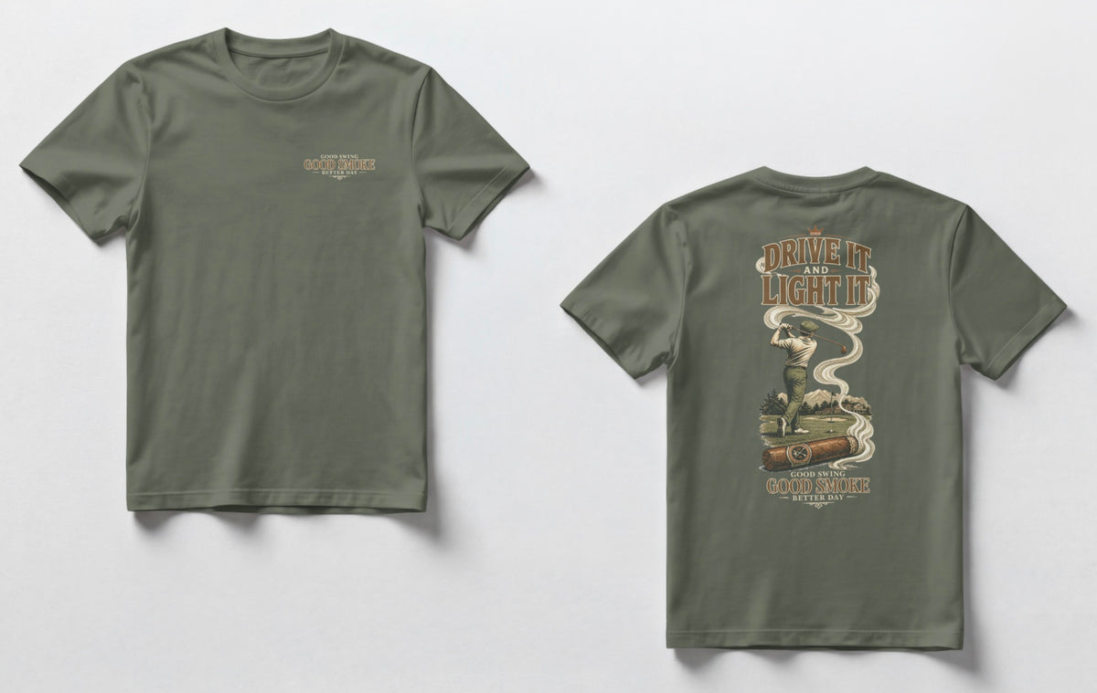 A military green t-shirt, front and back views. Front chest features small text reading 'Good Swing, Good Smoke, Better Day.' Back features a large vintage-style illustration of a golfer mid-swing on a course, with a cigar in the foreground releasing swirling smoke that wraps around the text 'Drive It and Light It' at the top, with 'Good Swing, Good Smoke, Better Day' printed below the graphic.