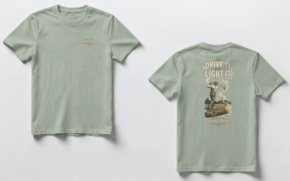 A sage green t-shirt, front and back views. Front chest features small text reading 'Good Swing, Good Smoke, Better Day.' Back features a large vintage-style illustration of a golfer mid-swing on a course, with a cigar in the foreground releasing swirling smoke that wraps around the text 'Drive It and Light It' at the top, with 'Good Swing, Good Smoke, Better Day' printed below the graphic.