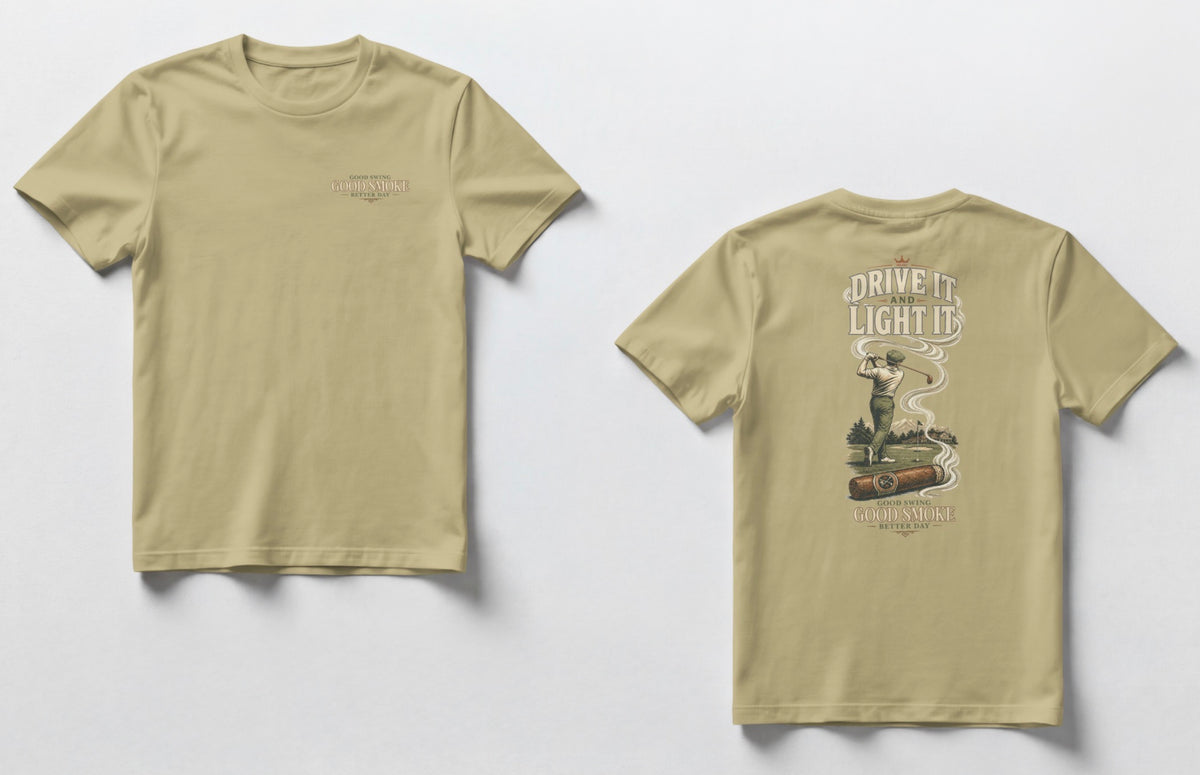 A sand color t-shirt, front and back views. Front chest features small text reading 'Good Swing, Good Smoke, Better Day.' Back features a large vintage-style illustration of a golfer mid-swing on a course, with a cigar in the foreground releasing swirling smoke that wraps around the text 'Drive It and Light It' at the top, with 'Good Swing, Good Smoke, Better Day' printed below the graphic.