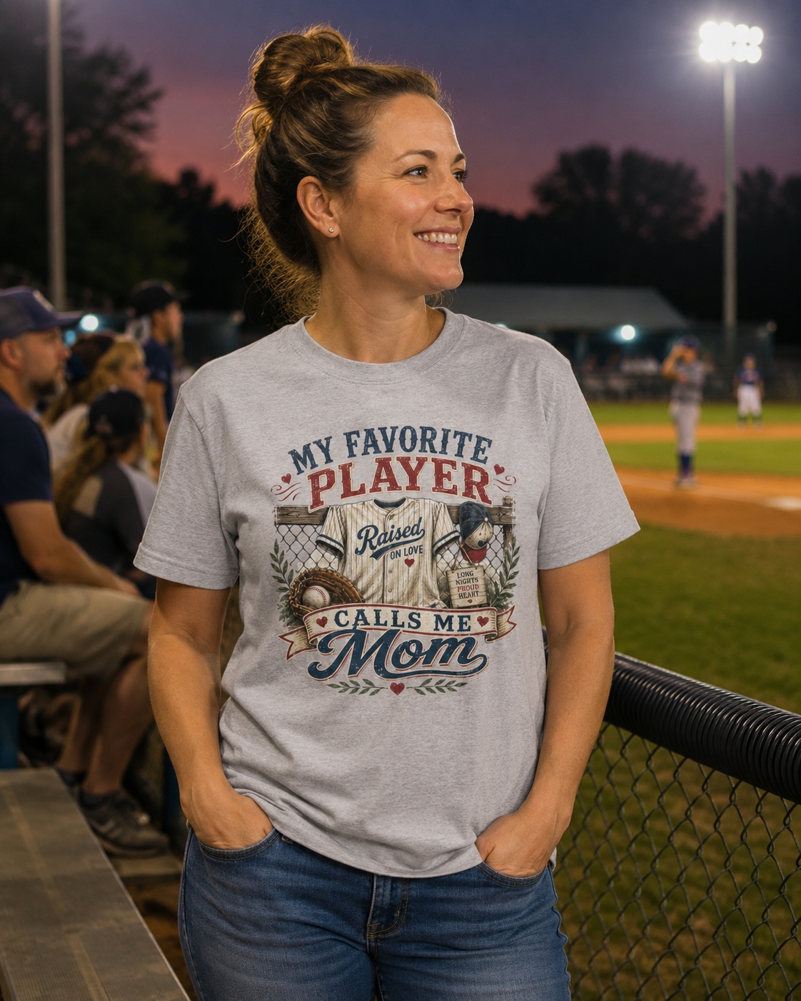A woman stands at a baseball field at dusk wearing a light gray graphic t-shirt that reads 'MY FAVORITE PLAYER CALLS ME Mom' with a baseball player illustration. The background shows a blurred baseball field and spectators.