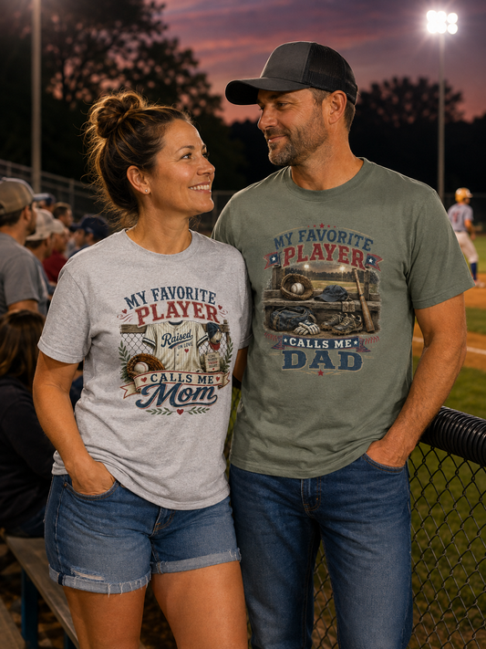 A man and a woman standing together on a baseball field at dusk, both wearing matching graphic t-shirts. The woman's light gray t-shirt reads 'MY FAVORITE PLAYER CALLS ME Mom', and the man's muted green t-shirt reads 'MY FAVORITE PLAYERS CALLS ME DAD'.