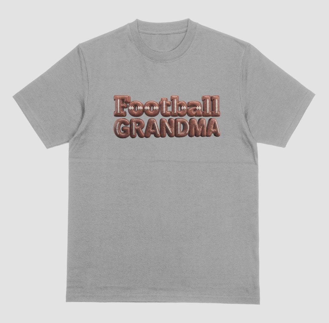 A grey crewneck t-shirt featuring the words 'Football GRANDMA' printed across the chest in a 3D block font filled with a brown football leather texture and white laces.