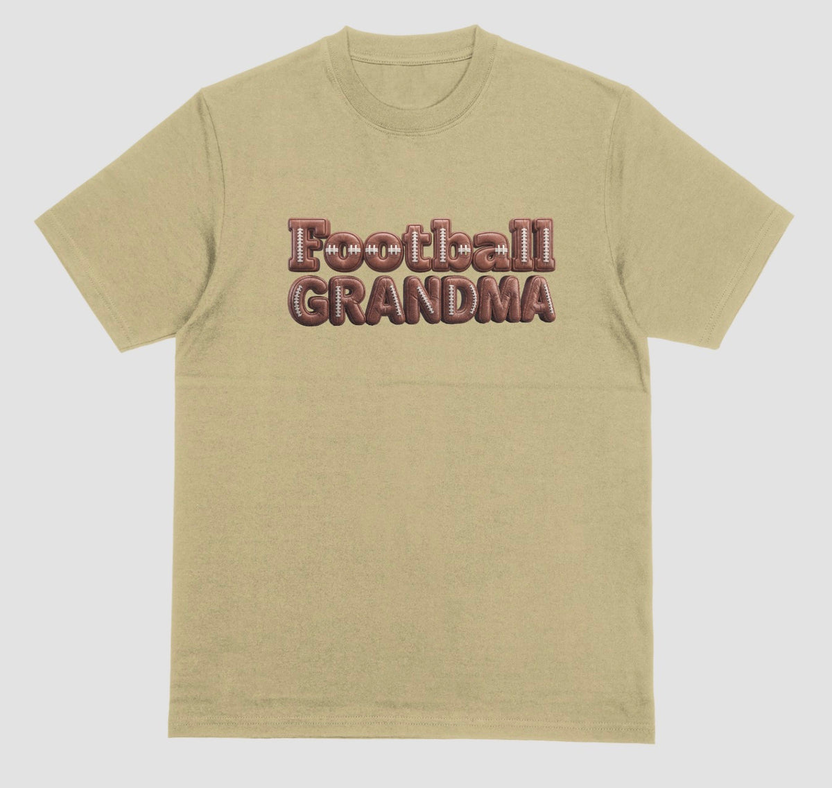 A sand color crewneck t-shirt featuring the words 'Football GRANDMA' printed across the chest in a 3D block font filled with a brown football leather texture and white laces.
