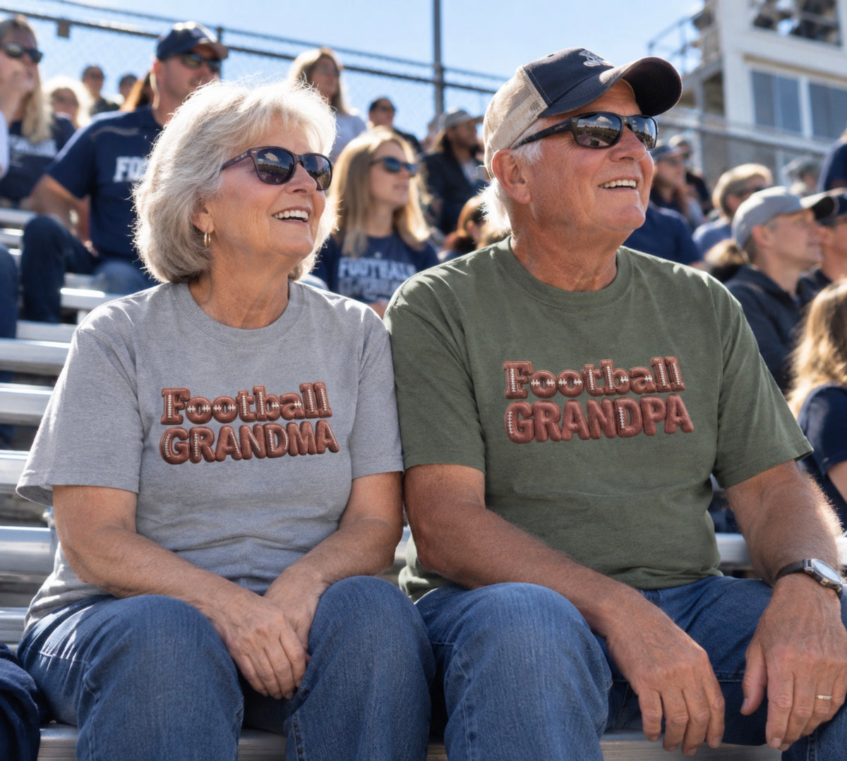 An grandma and grandpa sitting together in stadium bleachers, both wearing matching graphic T-shirts that read 'Football GRANDMA' and 'Football GRANDPA' in a 3D block font filled with a brown football leather texture and white laces. The man wears an military green shirt and a baseball cap, while the woman wears a heather gray shirt.