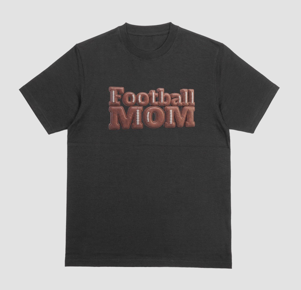 A black crewneck t-shirt featuring a 3D-style graphic on the chest that reads 'Football MOM' in a 3D block font filled with a brown football leather texture and white laces.