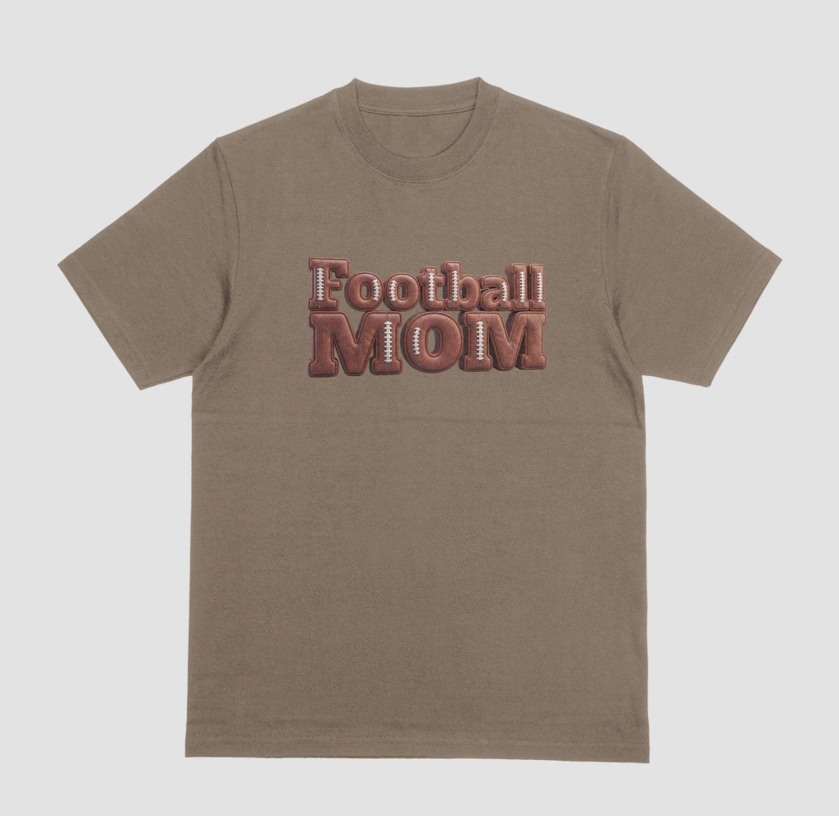 A brown savanna crewneck t-shirt featuring a 3D-style graphic on the chest that reads 'Football MOM' in a 3D block font filled with a brown football leather texture and white laces.