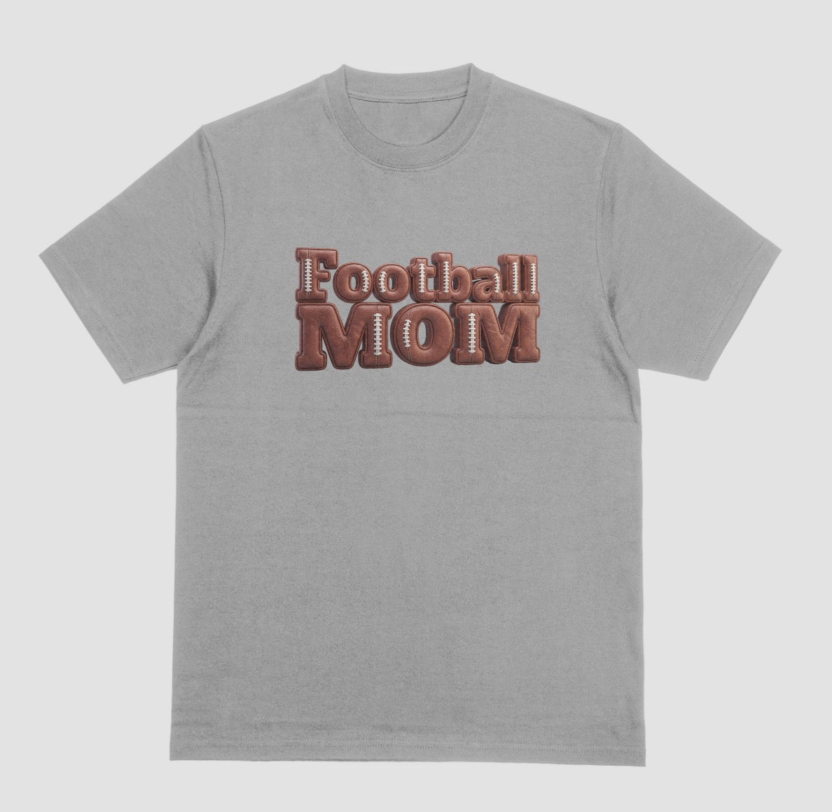A grey crewneck t-shirt featuring a 3D-style graphic on the chest that reads 'Football MOM' in a 3D block font filled with a brown football leather texture and white laces.