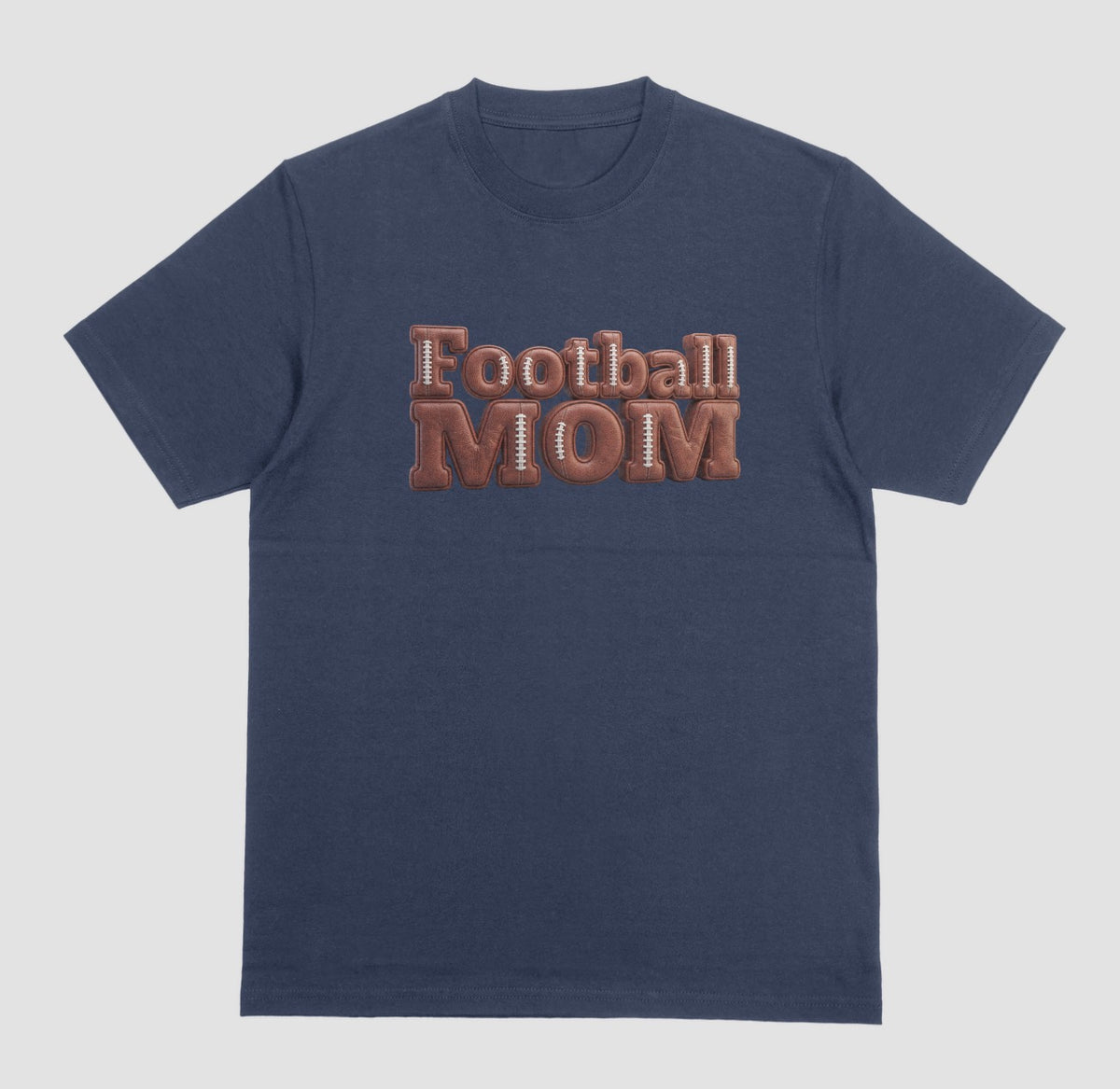 A navy blue crewneck t-shirt featuring a 3D-style graphic on the chest that reads 'Football MOM' in a 3D block font filled with a brown football leather texture and white laces.