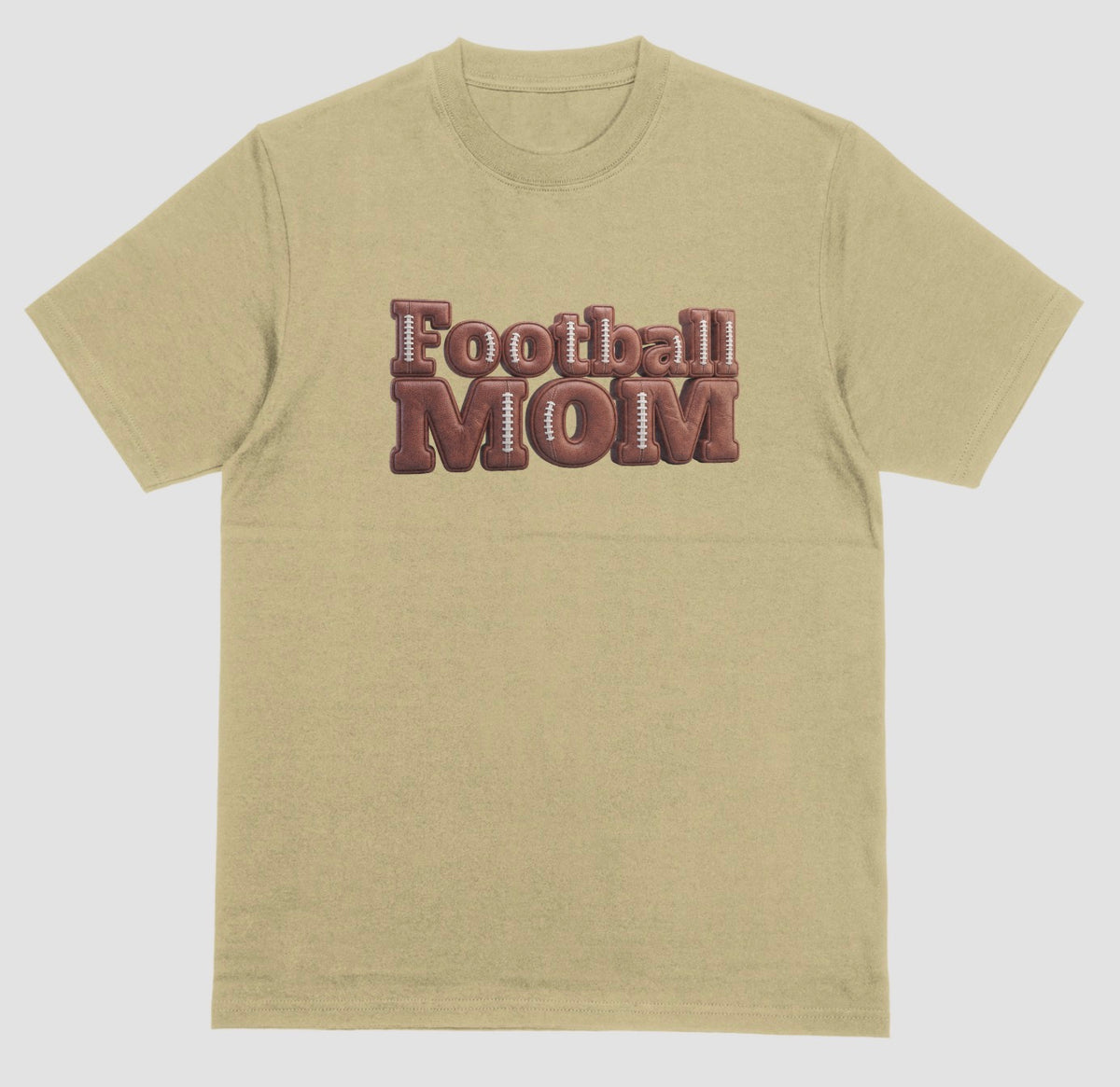 A sand color crewneck t-shirt featuring a 3D-style graphic on the chest that reads 'Football MOM' in a 3D block font filled with a brown football leather texture and white laces.