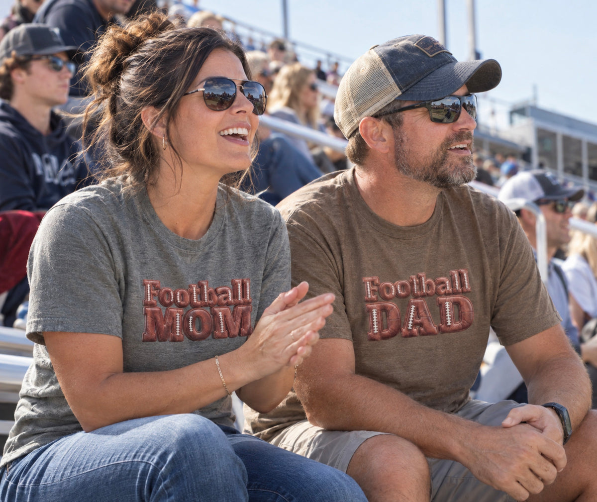 A man and woman sitting together in stadium seating at a football game, both wearing matching graphic T-shirts that read 'Football MOM' and 'Football DAD' in a 3D block font filled with a brown football leather texture and white laces.