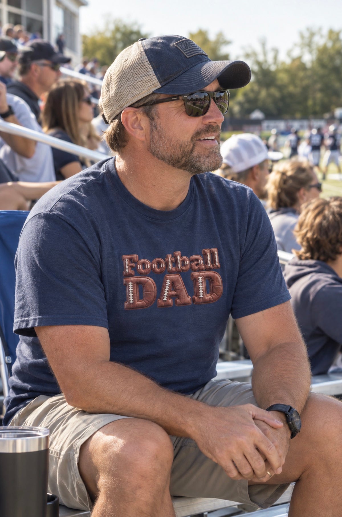 A man sitting in stadium seating at a sports event, wearing a navy blue t-shirt with 'Football DAD' printed on the front in a 3D block font filled with a brown football leather texture and white laces.