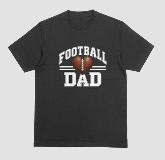 A black short-sleeve t-shirt with a 'FOOTBALL DAD' graphic on the chest. The graphic features a brown heart-shaped football in the center, flanked by two white horizontal stripes, with 'FOOTBALL' arched above and 'DAD' printed below in a distressed white font.