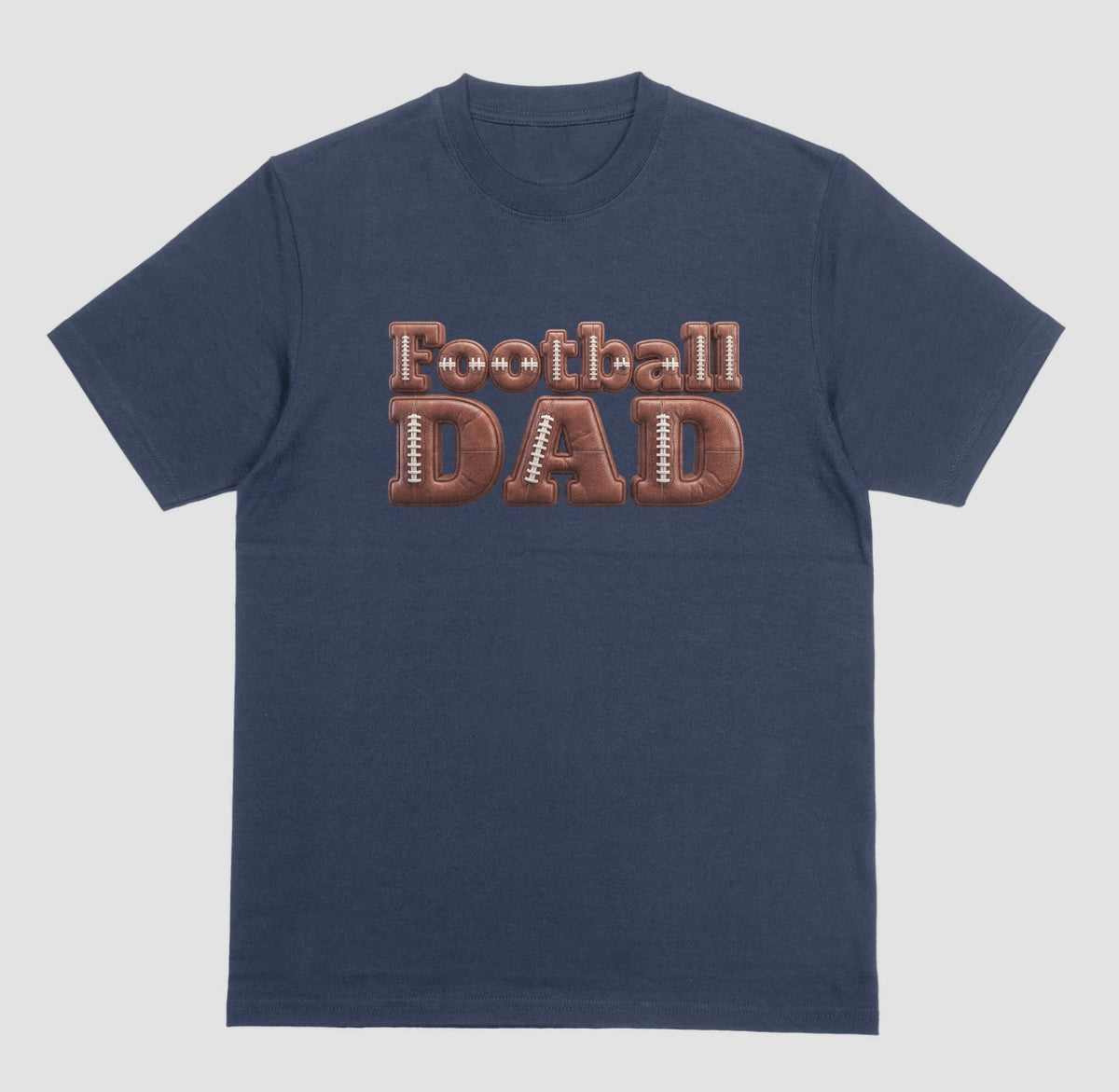 A navy blue crewneck t-shirt featuring a graphic on the chest that reads 'Football DAD' in a 3D block font filled with a brown football leather texture and white laces.