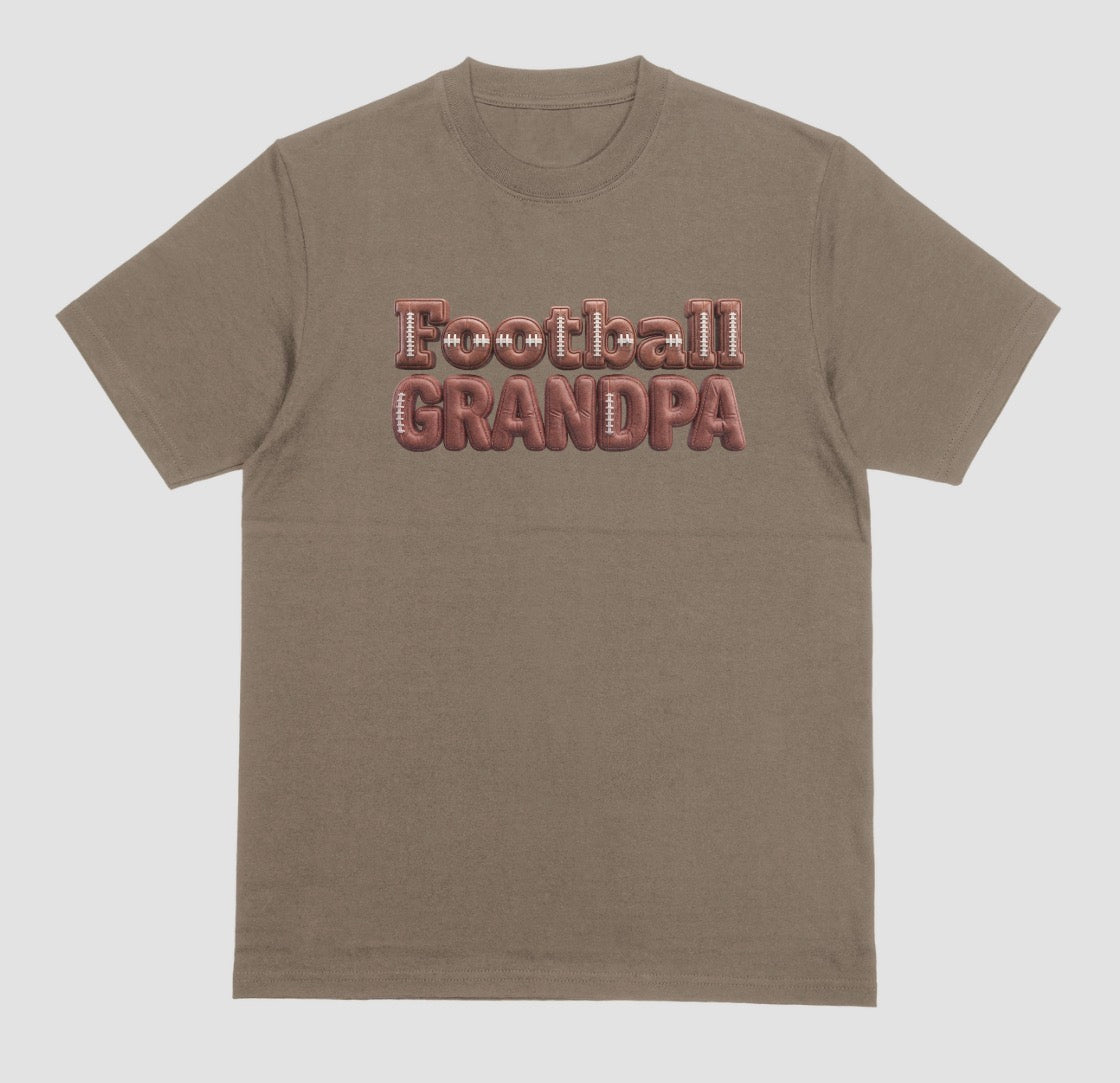 A brown savanna crewneck t-shirt featuring the words 'Football GRANDPA' printed across the chest in a 3D block font filled with a brown football leather texture and white laces.