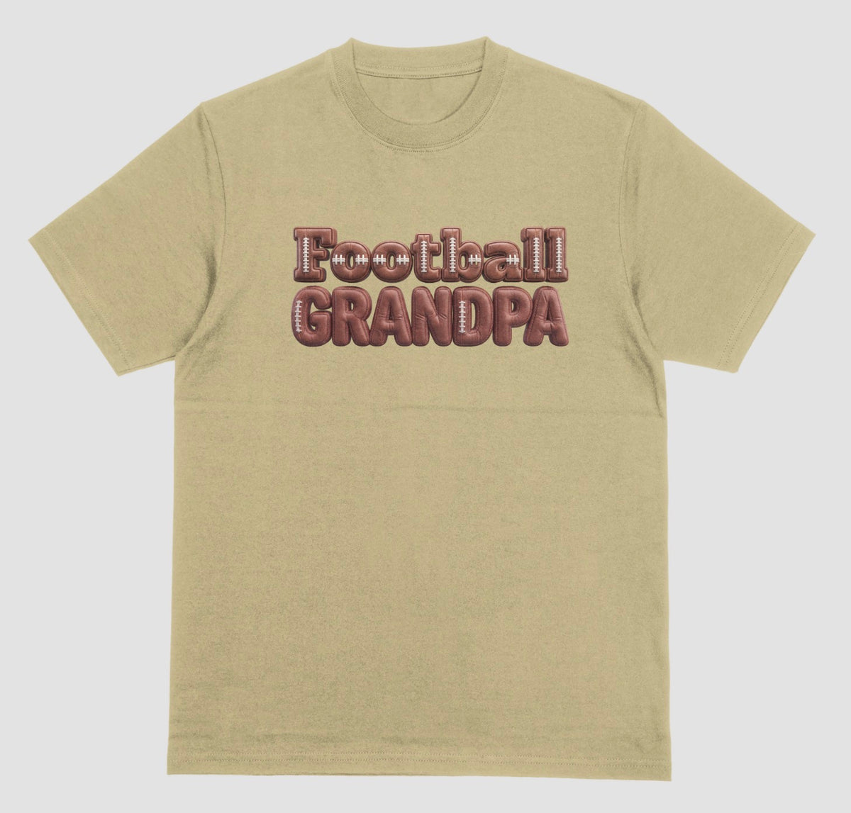A sand color crewneck t-shirt featuring the words 'Football GRANDPA' printed across the chest in a 3D block font filled with a brown football leather texture and white laces.