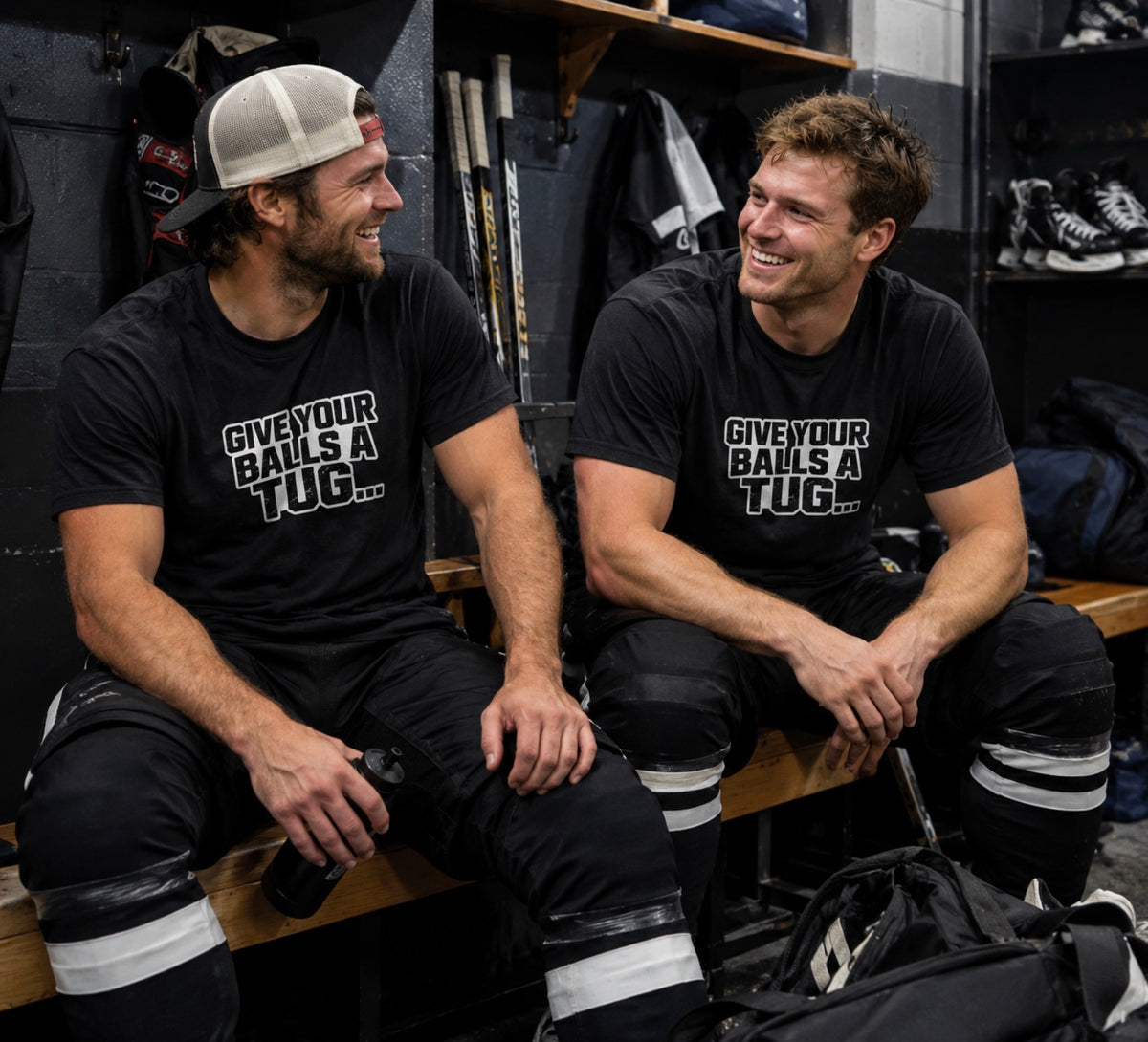 Two hockey players laughing in a locker room, both in black 'Give Your Balls a Tug...' tees with bold white block lettering.