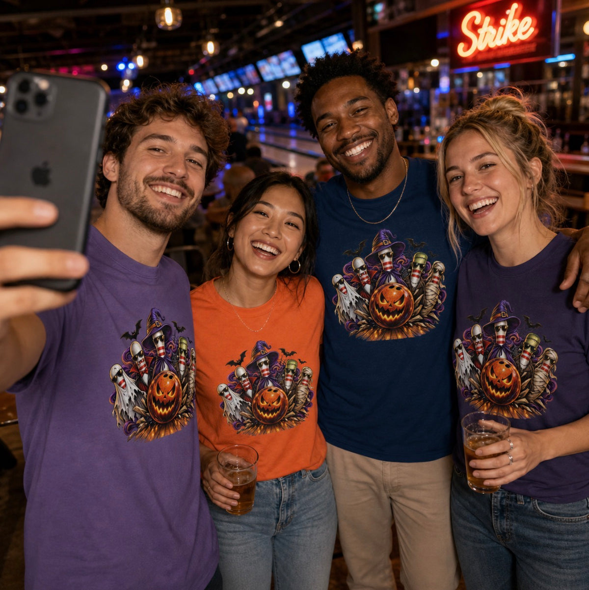 Four friends smiling for a selfie at a bowling alley bar, wearing matching Halloween bowling pin t-shirts in purple, orange, navy, and dark purple, two of them holding beers