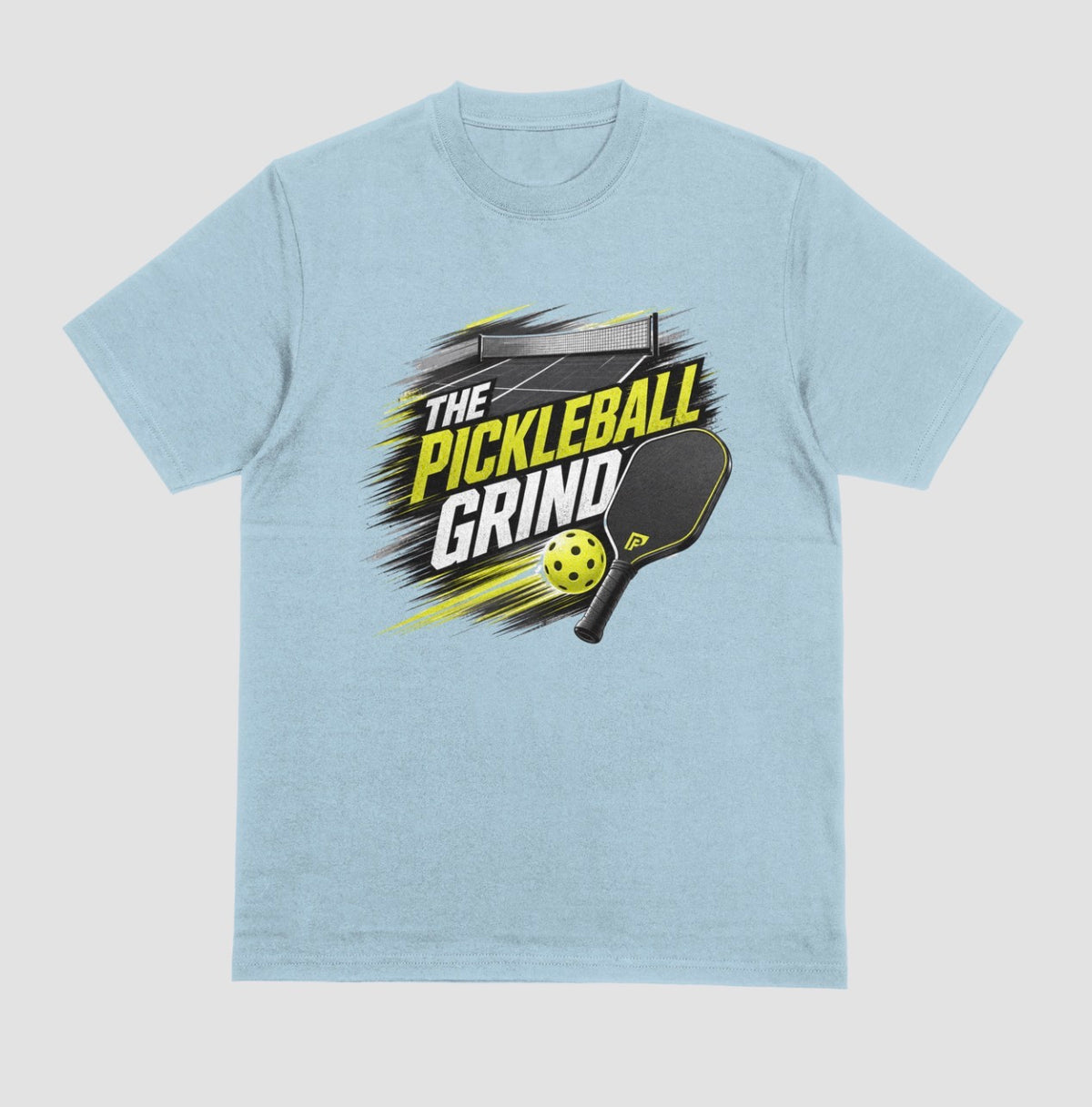 A light blue short-sleeve t-shirt with a 'The Pickleball Grind' graphic on the chest, featuring a yellow pickleball, black paddle, and yellow/black text over a gray brushstroke design, shown on a light gray background.