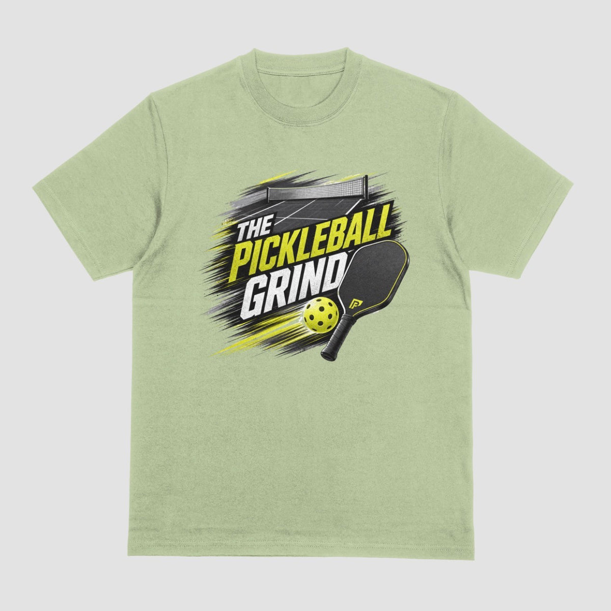 A pistachio green short-sleeve t-shirt with a 'The Pickleball Grind' graphic on the chest, featuring a yellow pickleball, black paddle, and yellow/black text over a gray brushstroke design, shown on a light gray background.
