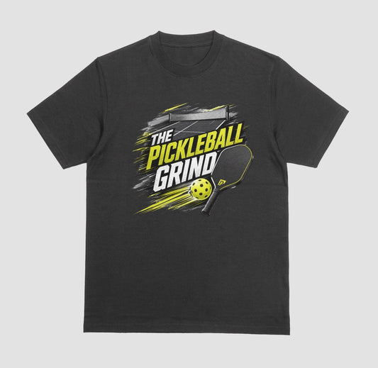 A black short-sleeve t-shirt with a 'The Pickleball Grind' graphic on the chest, featuring a yellow pickleball, black paddle, and yellow/black text over a gray brushstroke design, shown on a light gray background.
