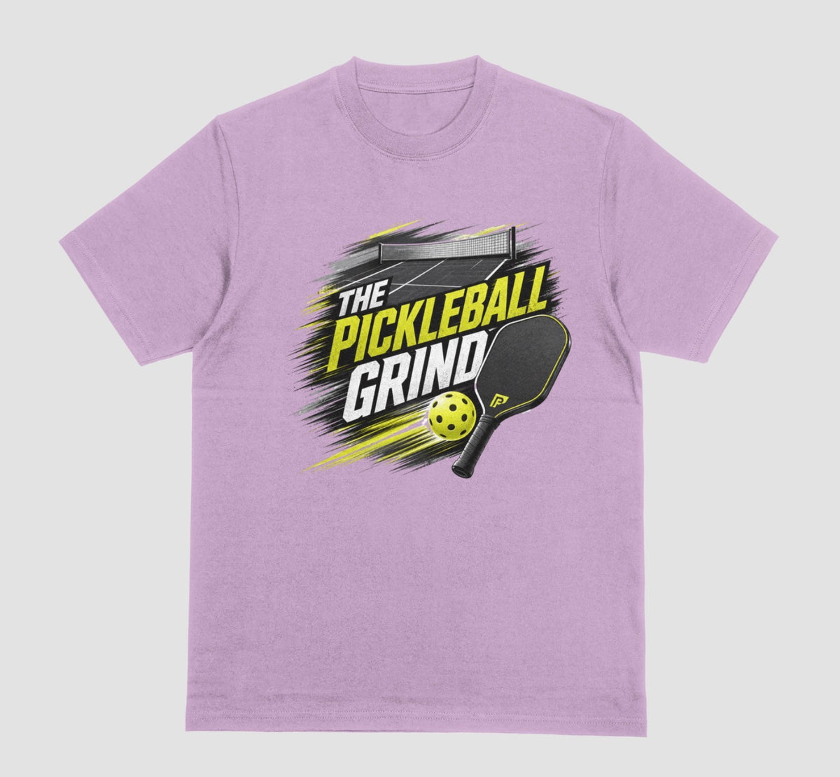 A lilac short-sleeve t-shirt with a 'The Pickleball Grind' graphic on the chest, featuring a yellow pickleball, black paddle, and yellow/black text over a gray brushstroke design, shown on a light gray background.