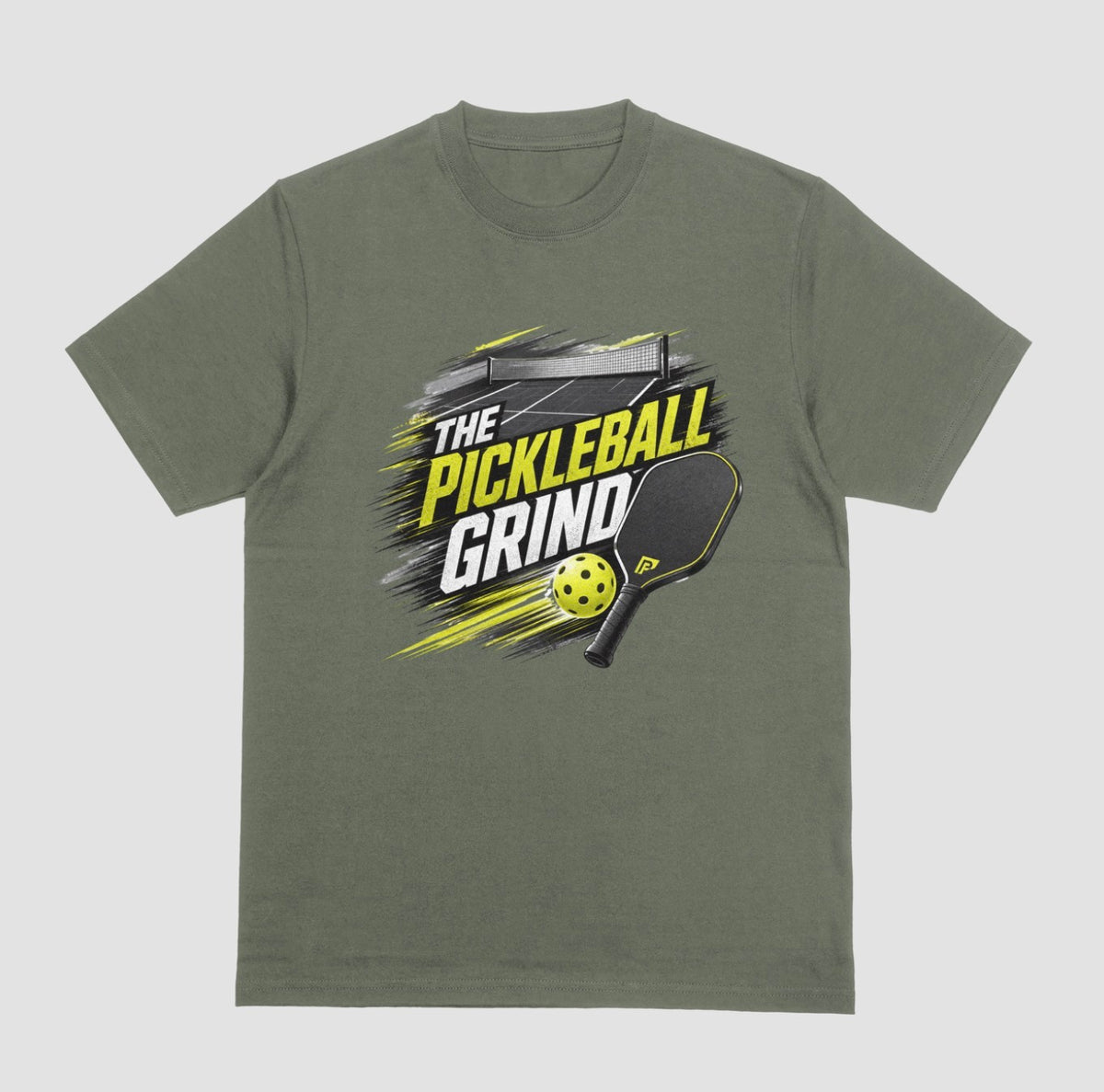 A military green short-sleeve t-shirt with a 'The Pickleball Grind' graphic on the chest, featuring a yellow pickleball, black paddle, and yellow/black text over a gray brushstroke design, shown on a light gray background.