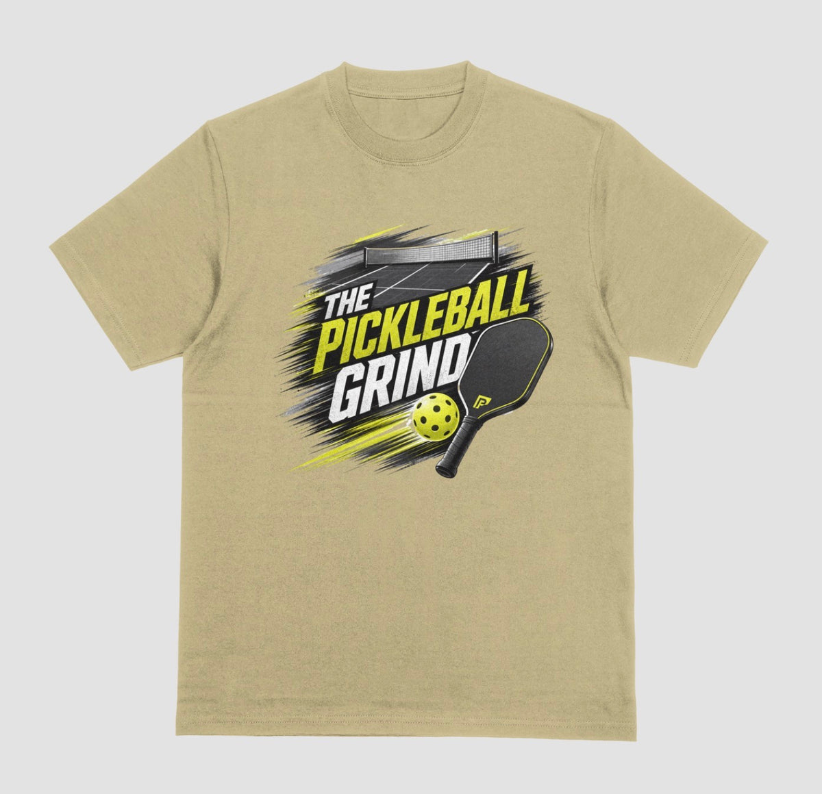 A sand color short-sleeve t-shirt with a 'The Pickleball Grind' graphic on the chest, featuring a yellow pickleball, black paddle, and yellow/black text over a gray brushstroke design, shown on a light gray background.
