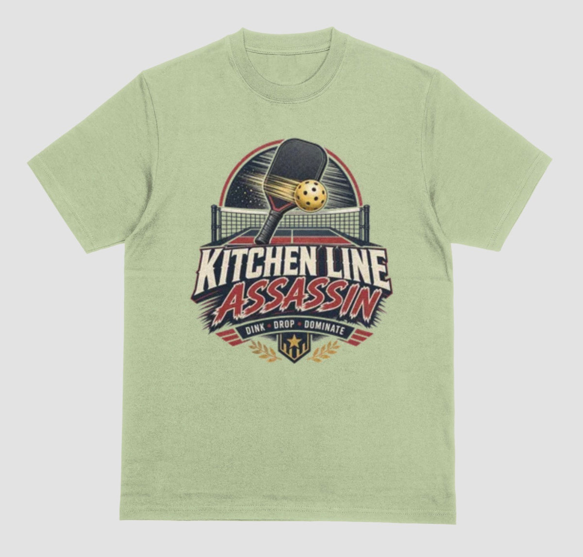 A pistachio green crewneck t-shirt featuring a graphic print on the front with a pickleball paddle and ball above the text 'KITCHEN LINE ASSASSIN' and 'DINK - DROP - DOMINATE'.