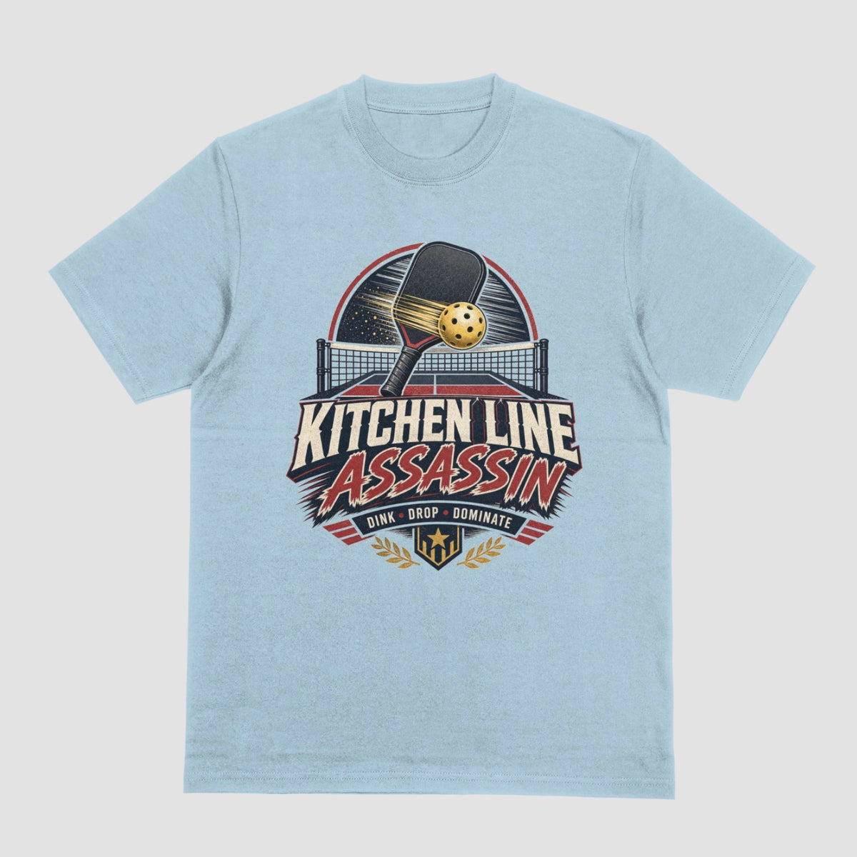 A light blue crewneck t-shirt featuring a graphic print on the front with a pickleball paddle and ball above the text 'KITCHEN LINE ASSASSIN' and 'DINK - DROP - DOMINATE'.